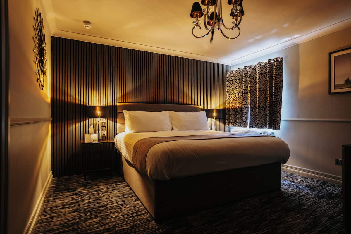 A hotel room with a large bed, a nightstand with a lamp, a wall-mounted light, and a window with patterned curtains.