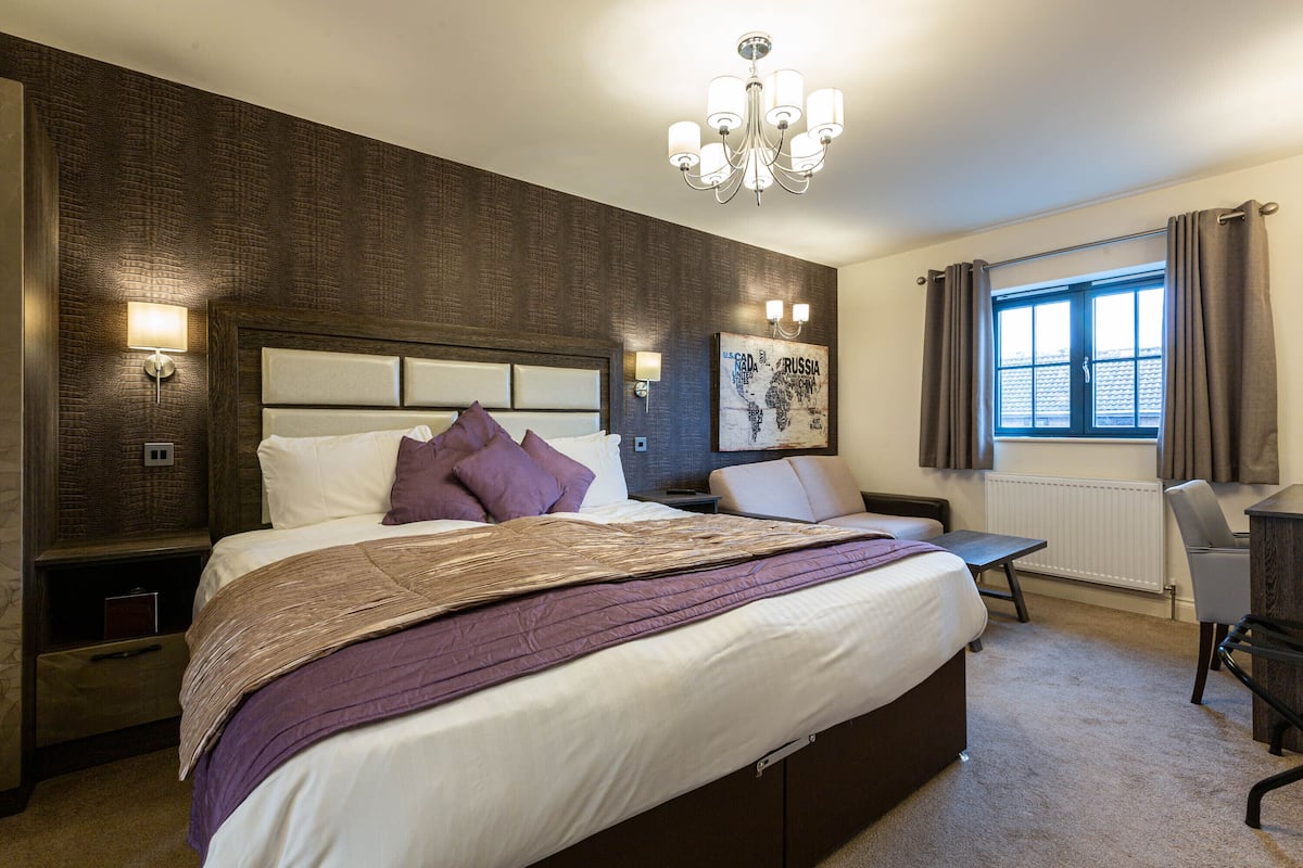A hotel room with a large bed, a desk, a chair, a chandelier, and a window with curtains.