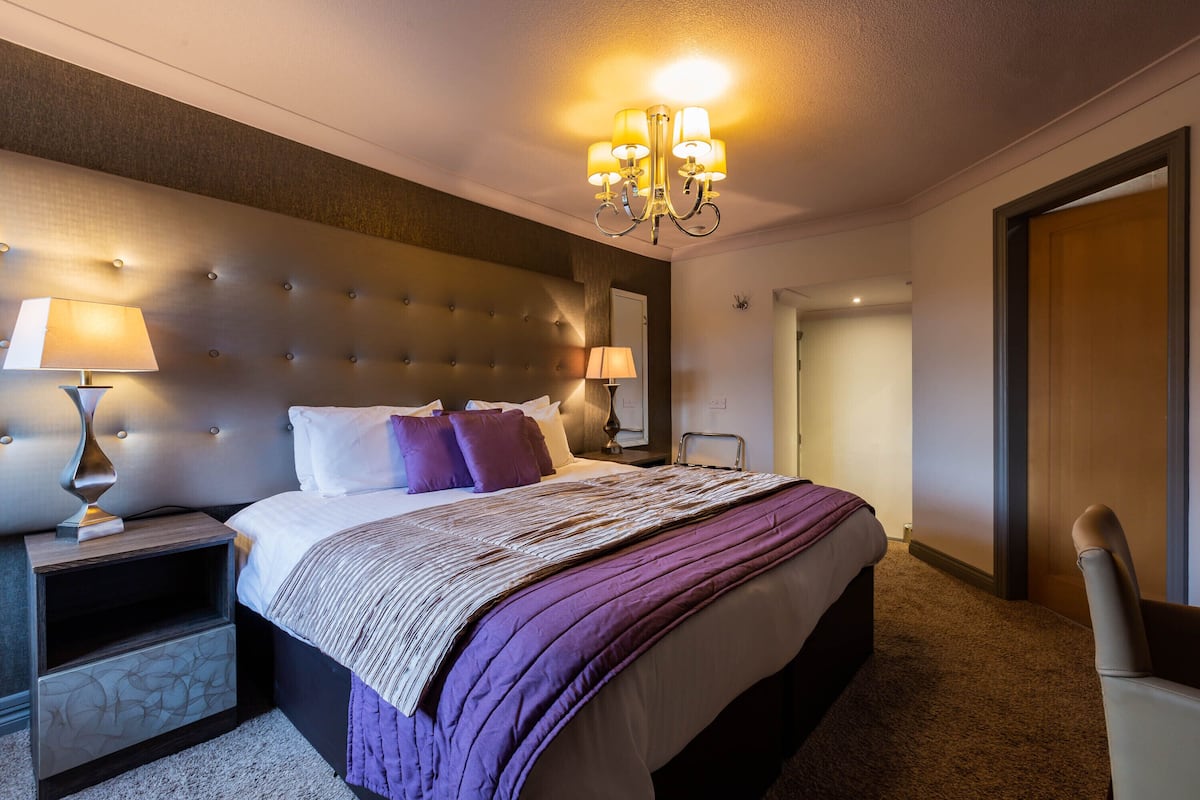 A modern hotel room with a tufted headboard, a large bed with a purple blanket, two bedside tables with lamps, and a chandelier.