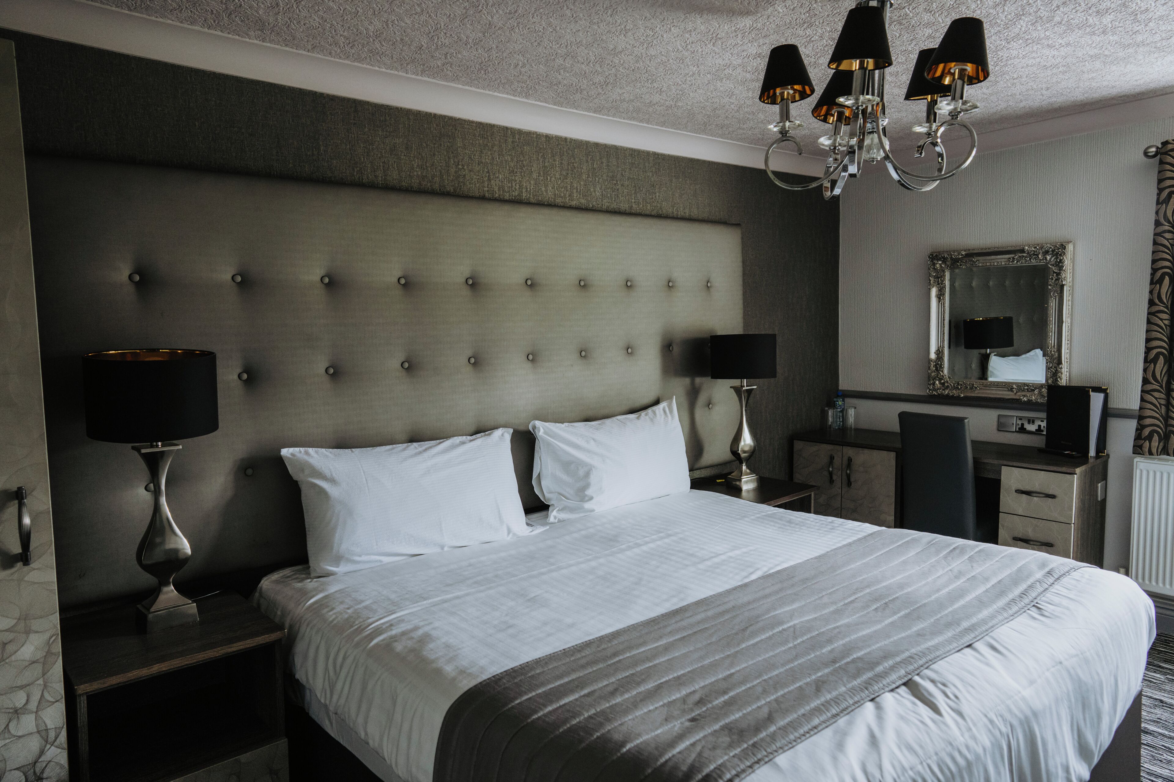 Una habitación de hotel con una cama grande, dos mesitas de noche con lámparas, un aparador, un espejo y una araña.