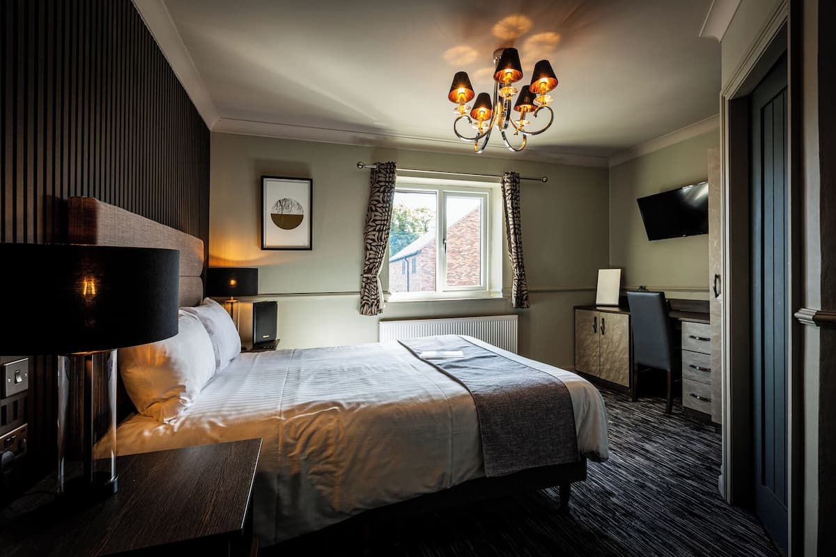A hotel room with a bed, bedside lamp, window with curtains, radiator, desk, chair, and a TV mounted on the wall.