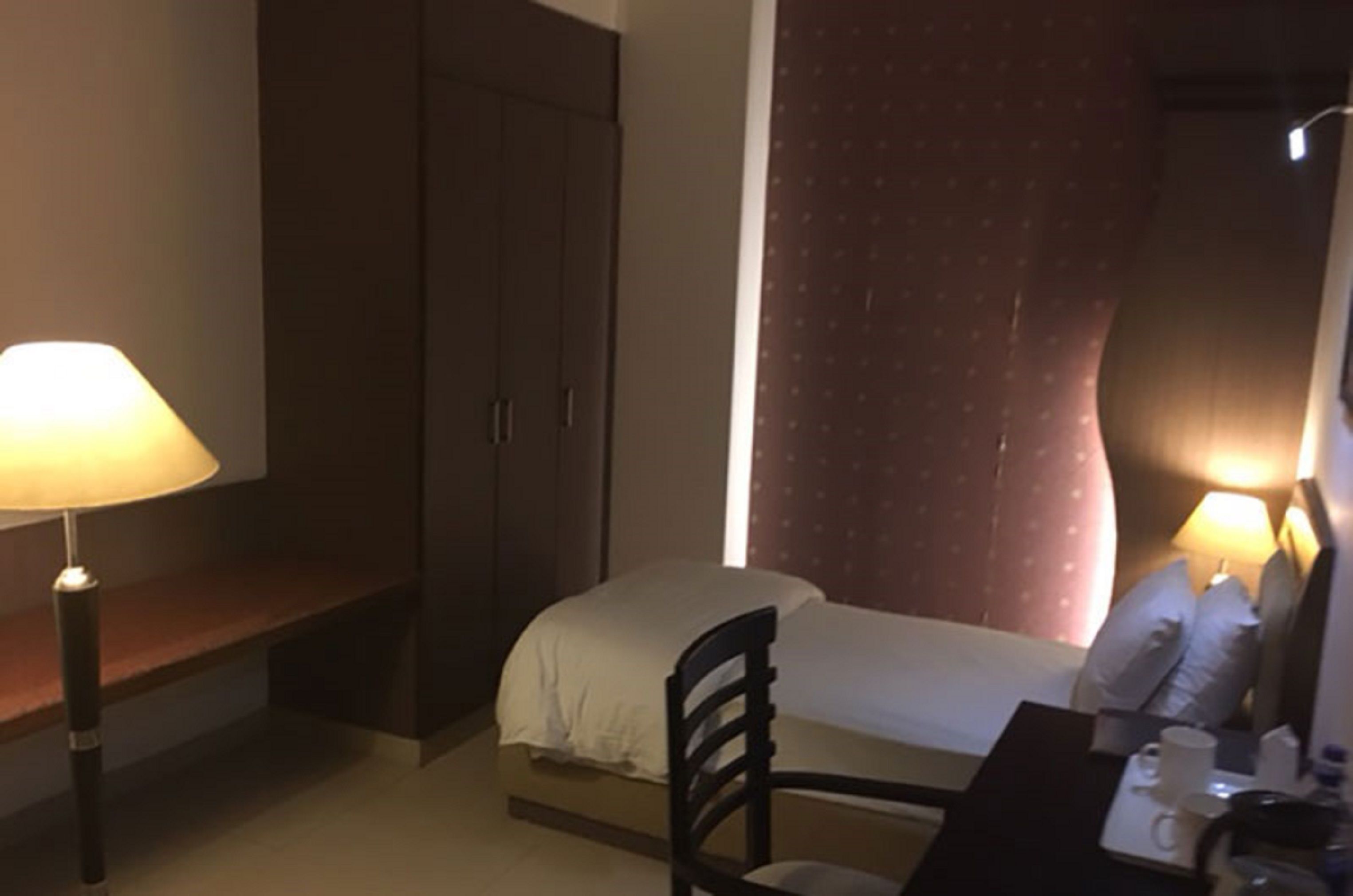 A hotel room with a bed, a chair, a desk with a lamp, and a wardrobe.