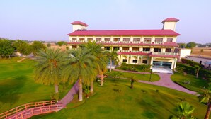 A large, multi-story hotel with a red roof and white walls, surrounded by well-maintained lawns and palm trees. - Shiva Oasis Resort (Behror)