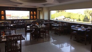 A spacious restaurant with multiple tables and chairs, large windows offering a view of greenery, and a modern ceiling design. - Shiva Oasis Resort (Behror)