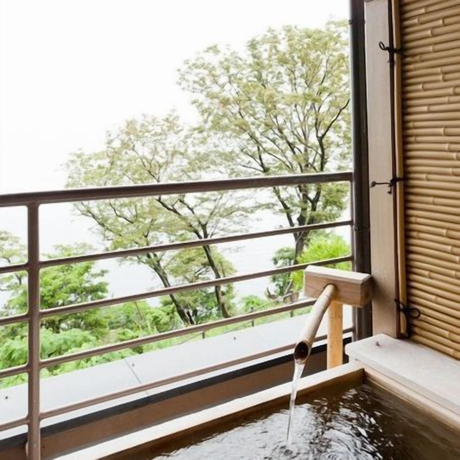 A hot spring bath with a wooden faucet, a view of trees through a window, and a bamboo blind.