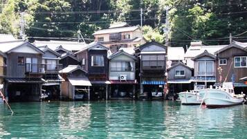 A row of traditional wooden houses with a waterway in front, boats docked, and a forested hillside in the background.