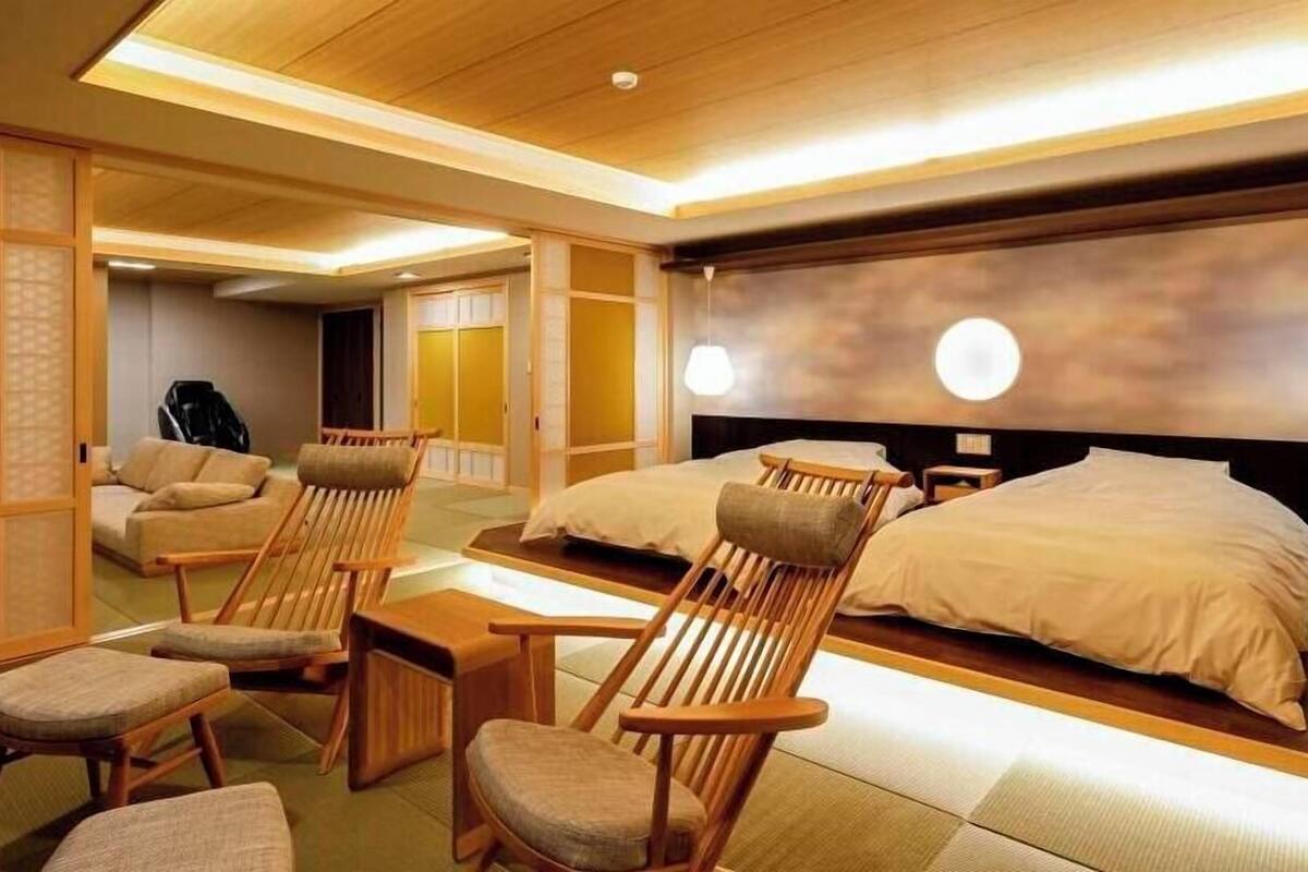 A traditional Japanese-style hotel room with tatami flooring, a low wooden bed, a futon bed, and a seating area with wooden chairs and a table.