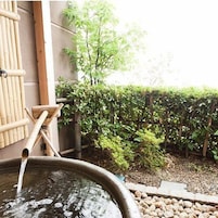Un onsen japonés tradicional con un baño de piedra, estructuras de bambú y un jardín con valla de madera y follaje.