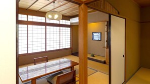 Une pièce de style japonais traditionnel, avec un sol recouvert de tatamis, une petite table basse avec des coussins, un luminaire suspendu et une porte coulissante donnant sur une autre pièce.