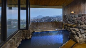 Un bain thermal en plein air, avec une terrasse en bois, des murs en pierre et une vue sur le paysage urbain et les montagnes.