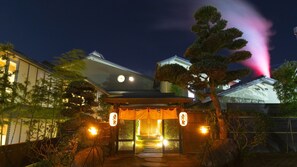 Un bâtiment japonais traditionnel, doté d’une entrée couverte, d’un chemin de pierre menant à la porte et d’un arbre en premier plan.