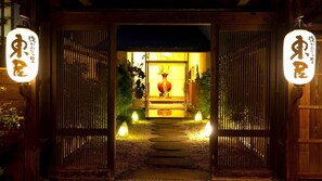 Une porte d’entrée traditionnelle japonaise, agrémentée d’enseignes illuminées, qui mène à un espace intérieur chaleureux, éclairé d’une lumière douce, et où trône une lanterne rouge au centre.