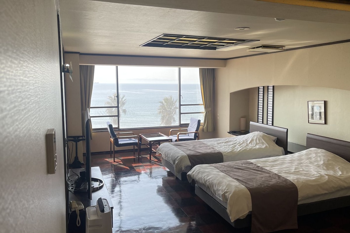 A hotel room with two beds, a desk, a chair, and a large window with a view of the ocean.