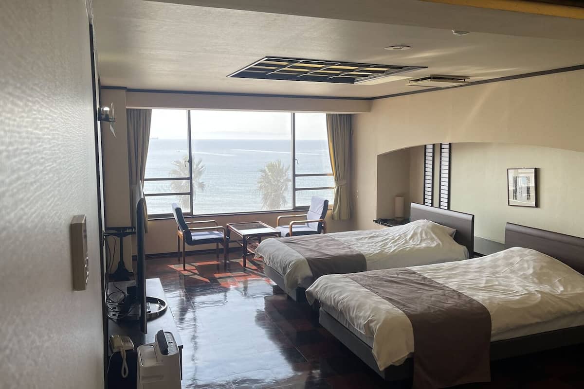 A hotel room with two beds, a desk, a chair, and a large window with a view of the ocean.