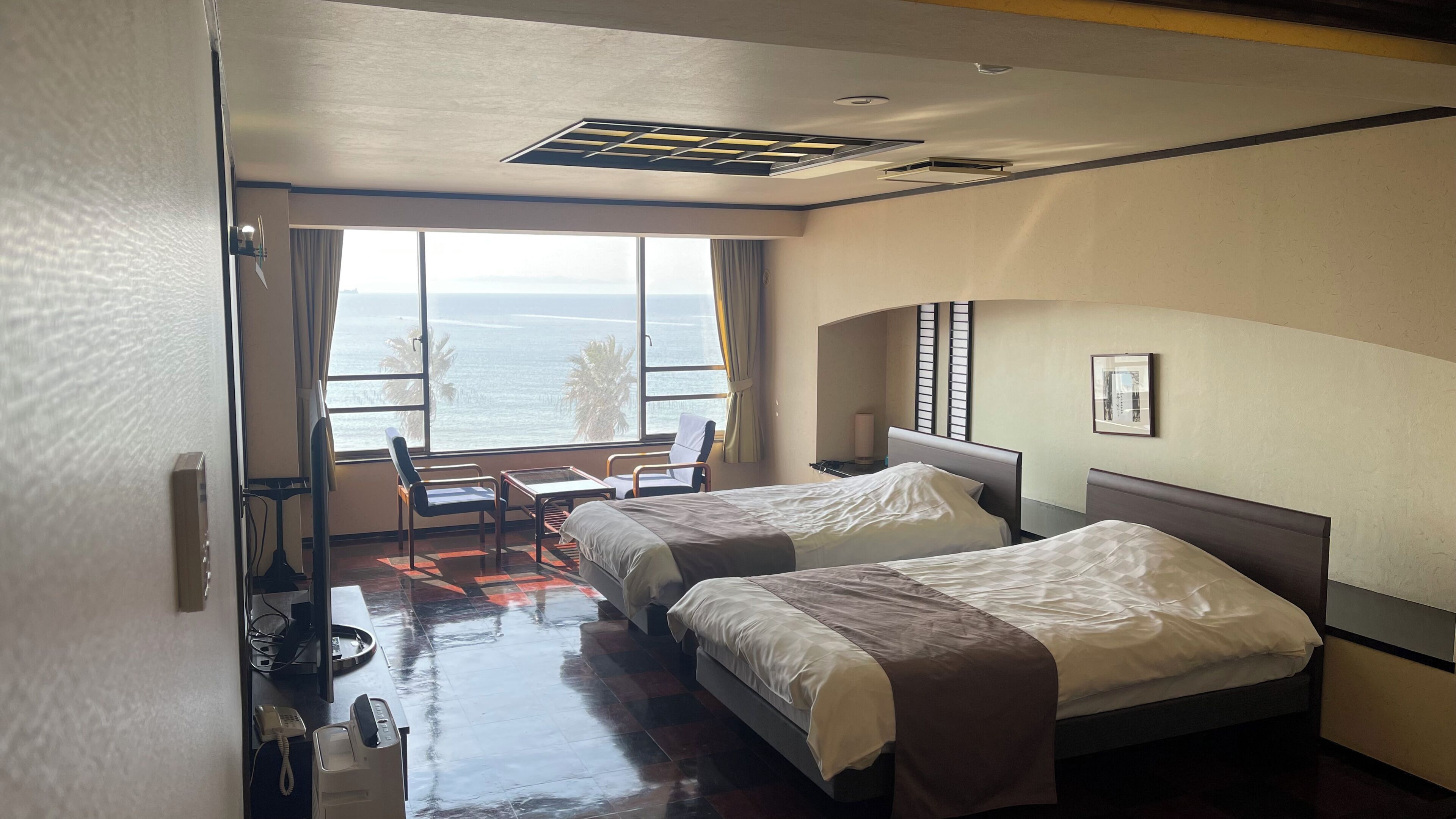 Une chambre d’hôtel avec deux lits, un bureau, une chaise et une grande fenêtre donnant sur la mer.