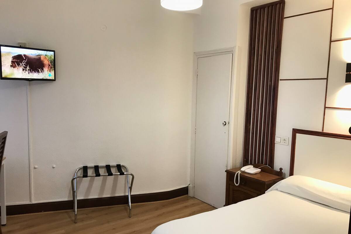 A hotel room with a bed, a bedside table with a phone, a small table with a chair, a TV mounted on the wall, and a door.