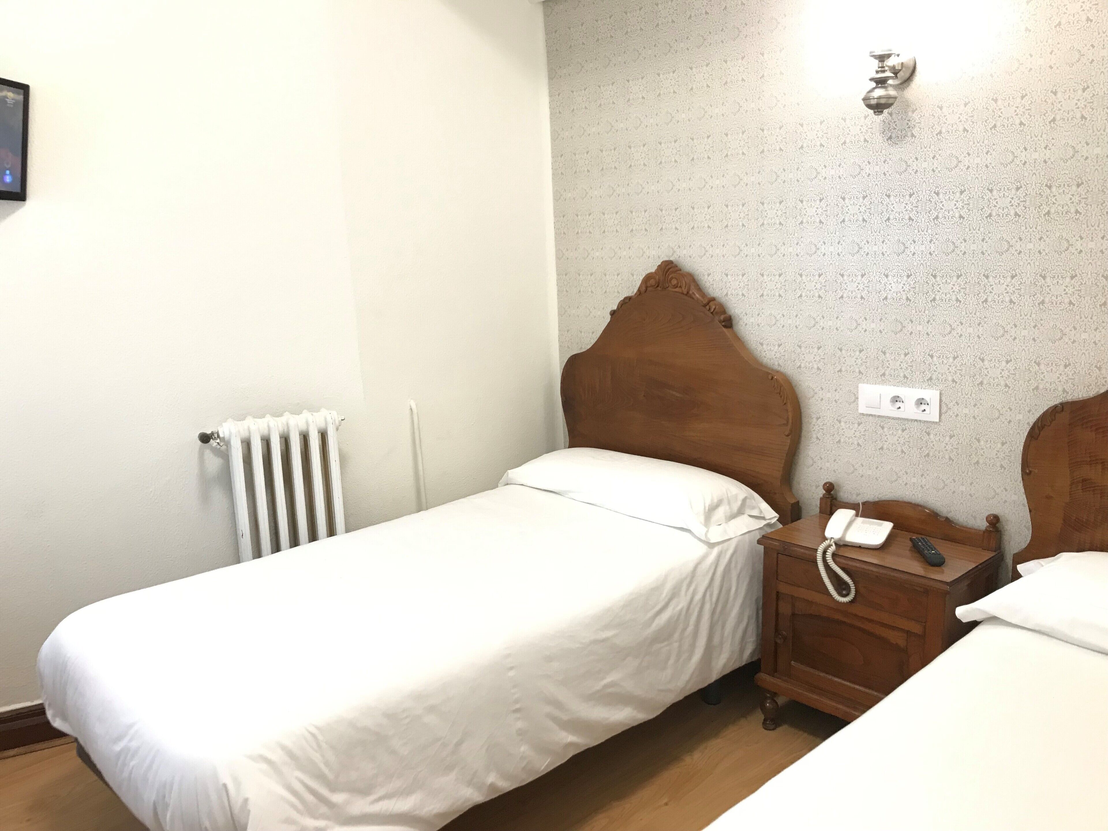 Een kleine eenpersoonskamer met een houten hoofdbord, een nachtkastje met telefoon en een radiator aan de muur.