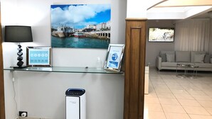 A reception area with a glass desk displaying informational posters, a digital screen, a standing heater, and a sofa in the background. - Hostal La Mexicana (Santander)