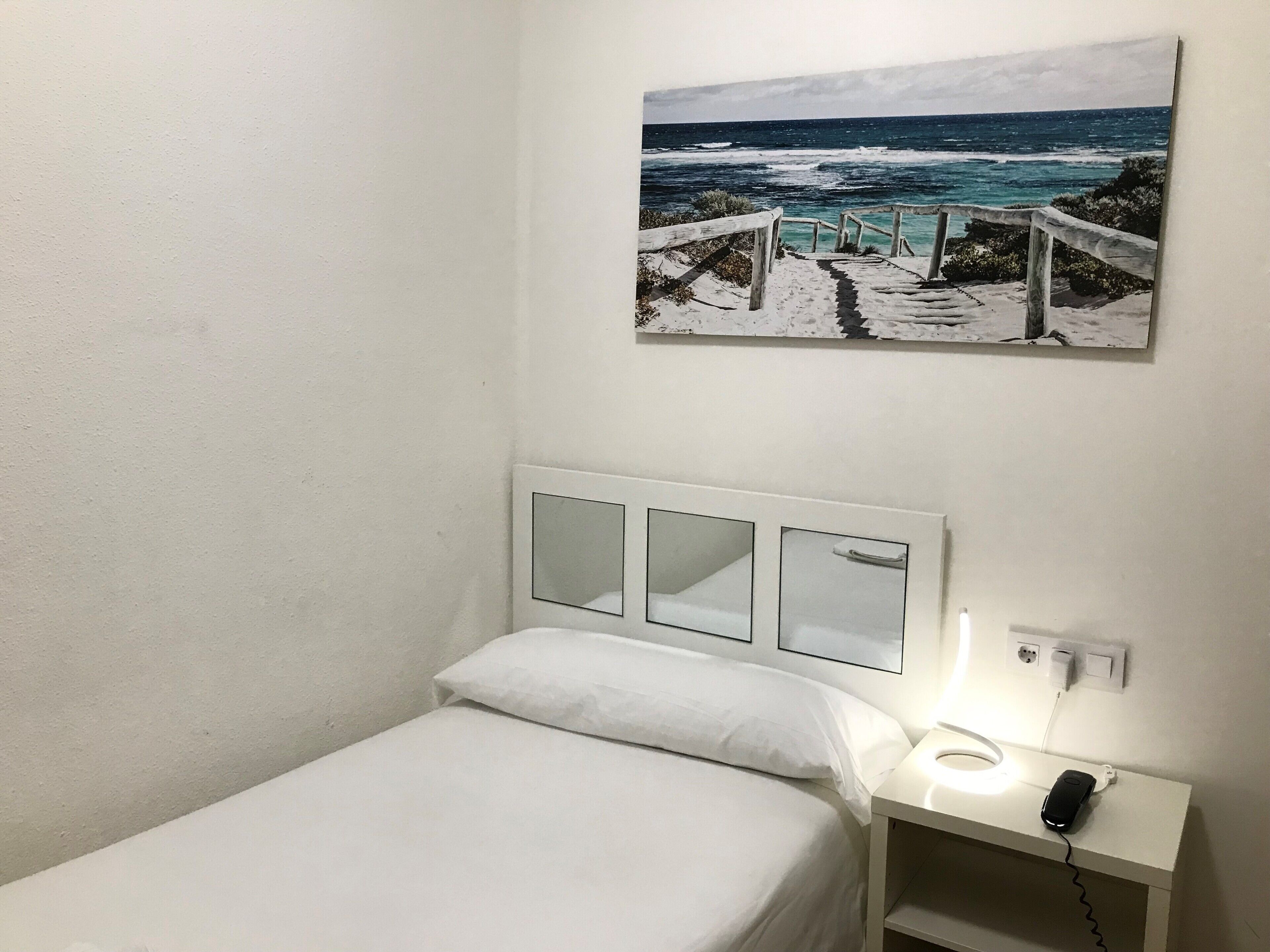 Una cama individual con ropa de cama blanca, una mesita de noche con lámpara y un cuadro de una escena costera.