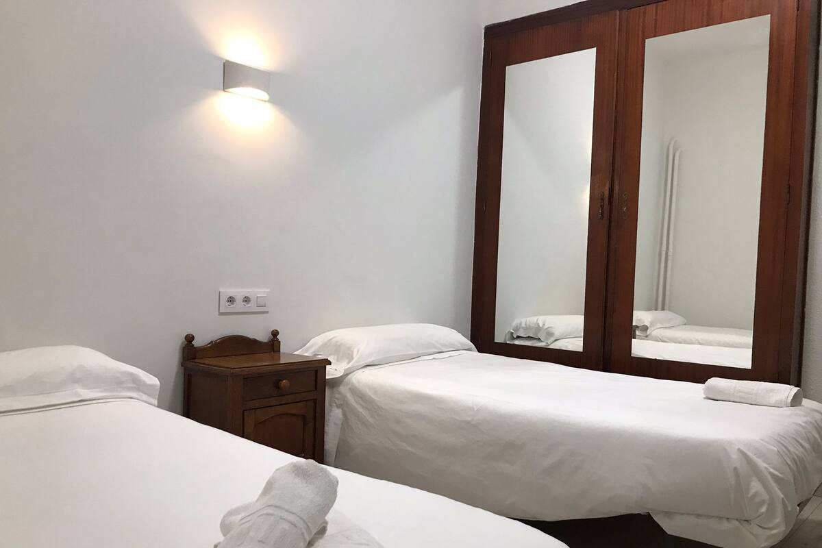 A hotel room with two single beds, a wooden nightstand, a wall-mounted light, and a large mirror.
