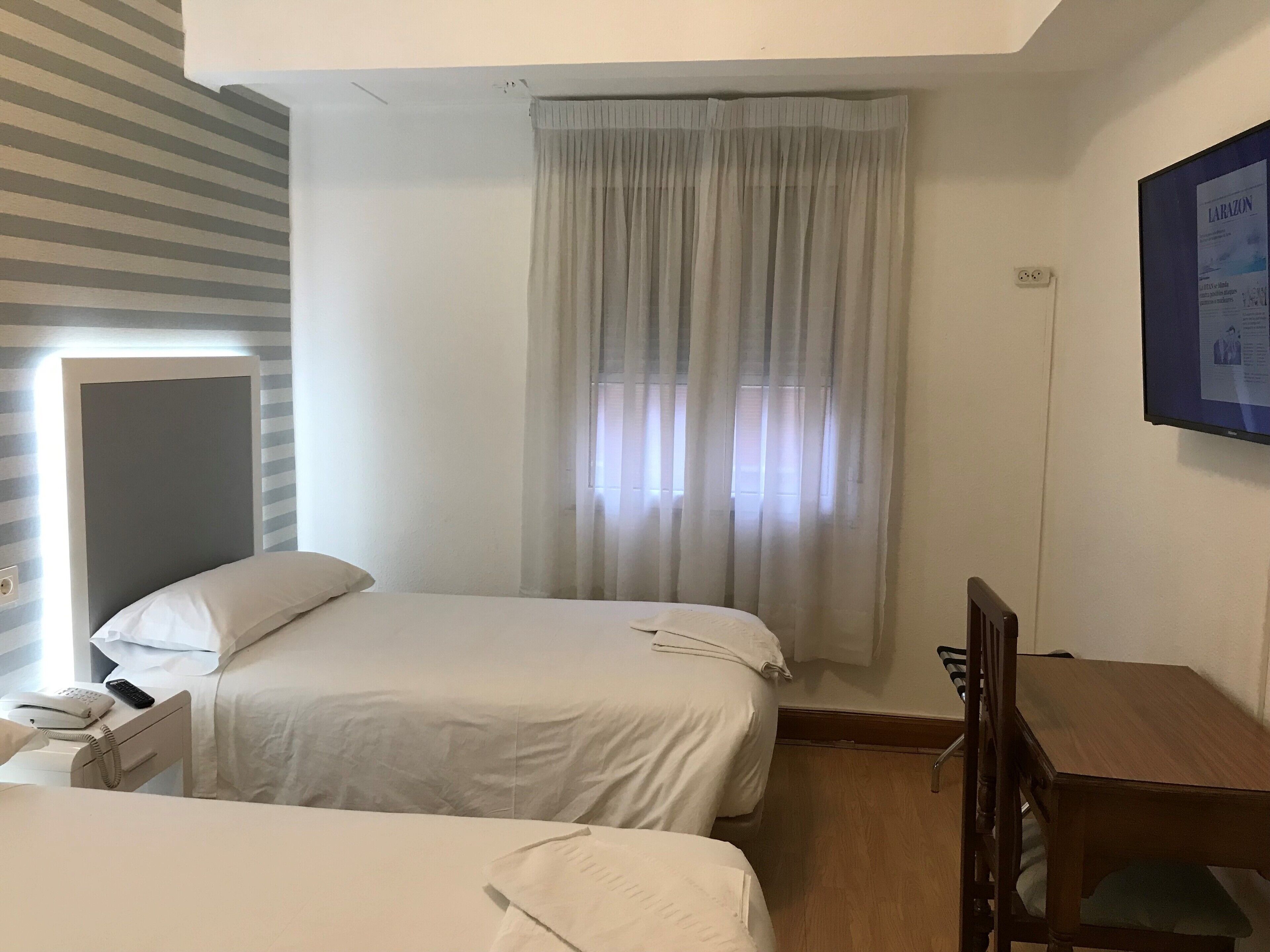 Una habitación de hotel con dos camas, un escritorio de madera y un ventanal con cortinas translúcidas.