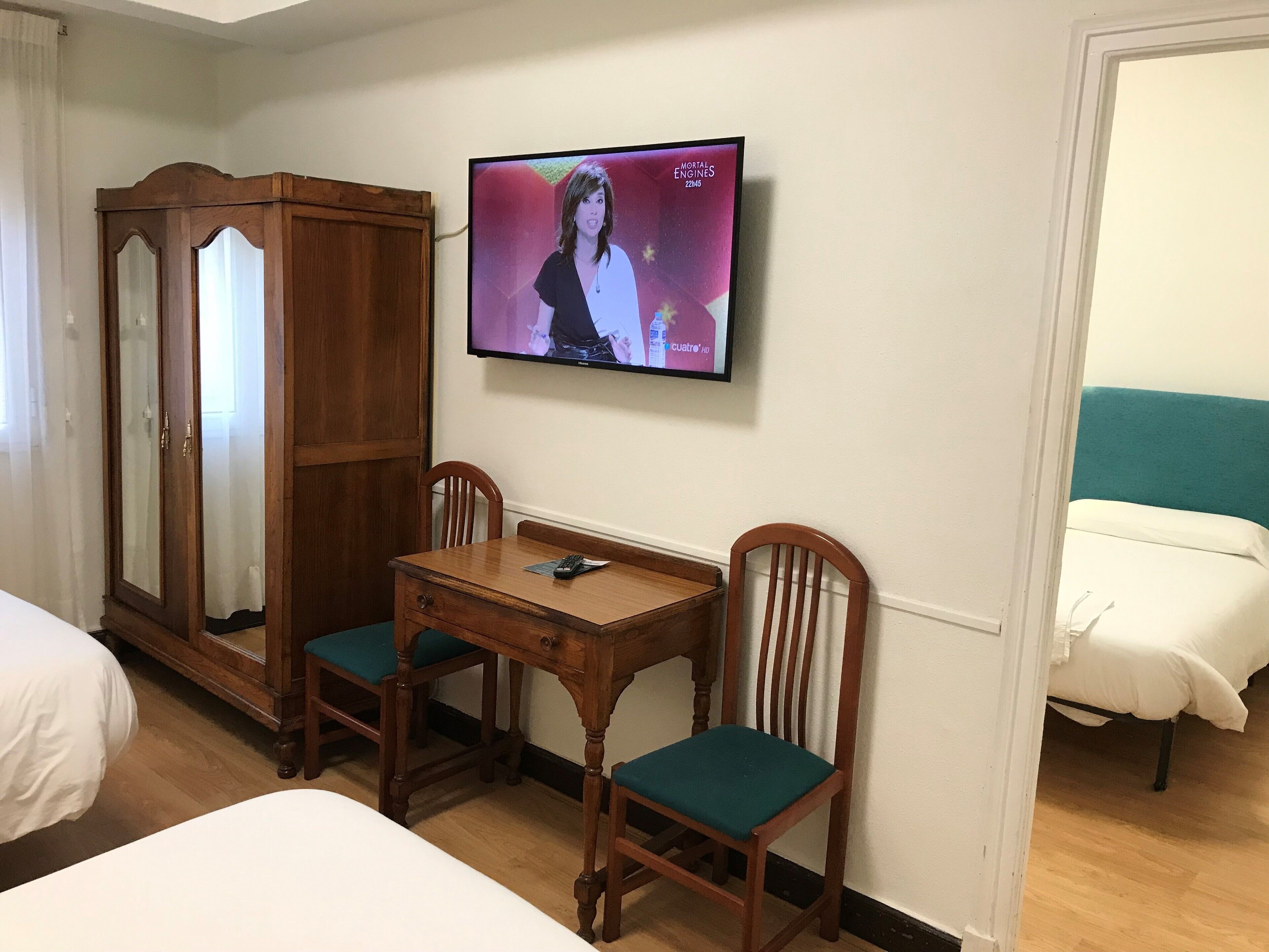 A hotel room with a wooden cabinet, a TV mounted on the wall, a wooden table with chairs, and two beds.