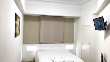 A hotel room with a bed, a flat-screen TV mounted on the wall, a window with a blind, and a painting on the wall.