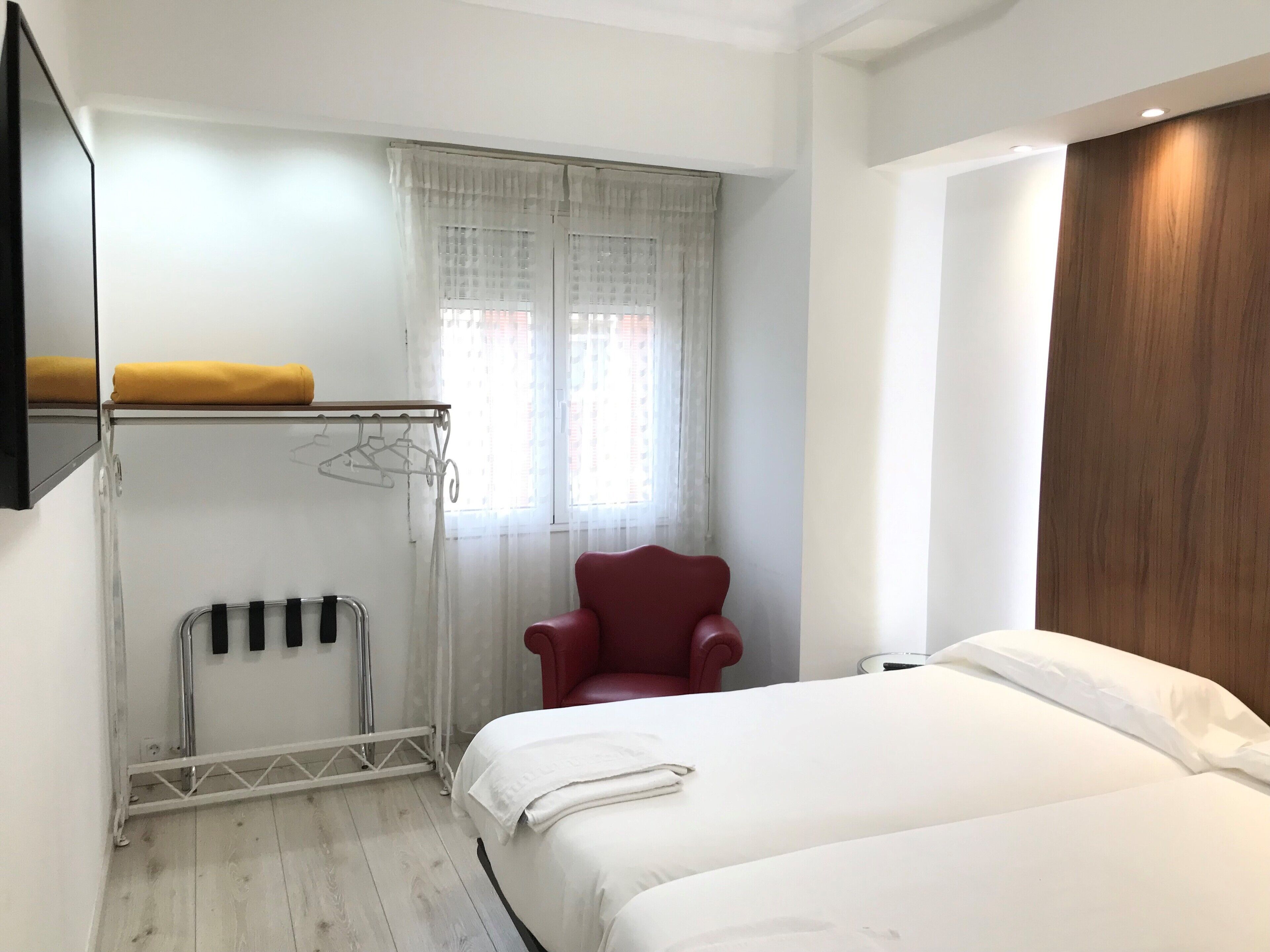 A hotel room with a bed, a red chair, a coat rack with hangers, and a window with sheer curtains.
