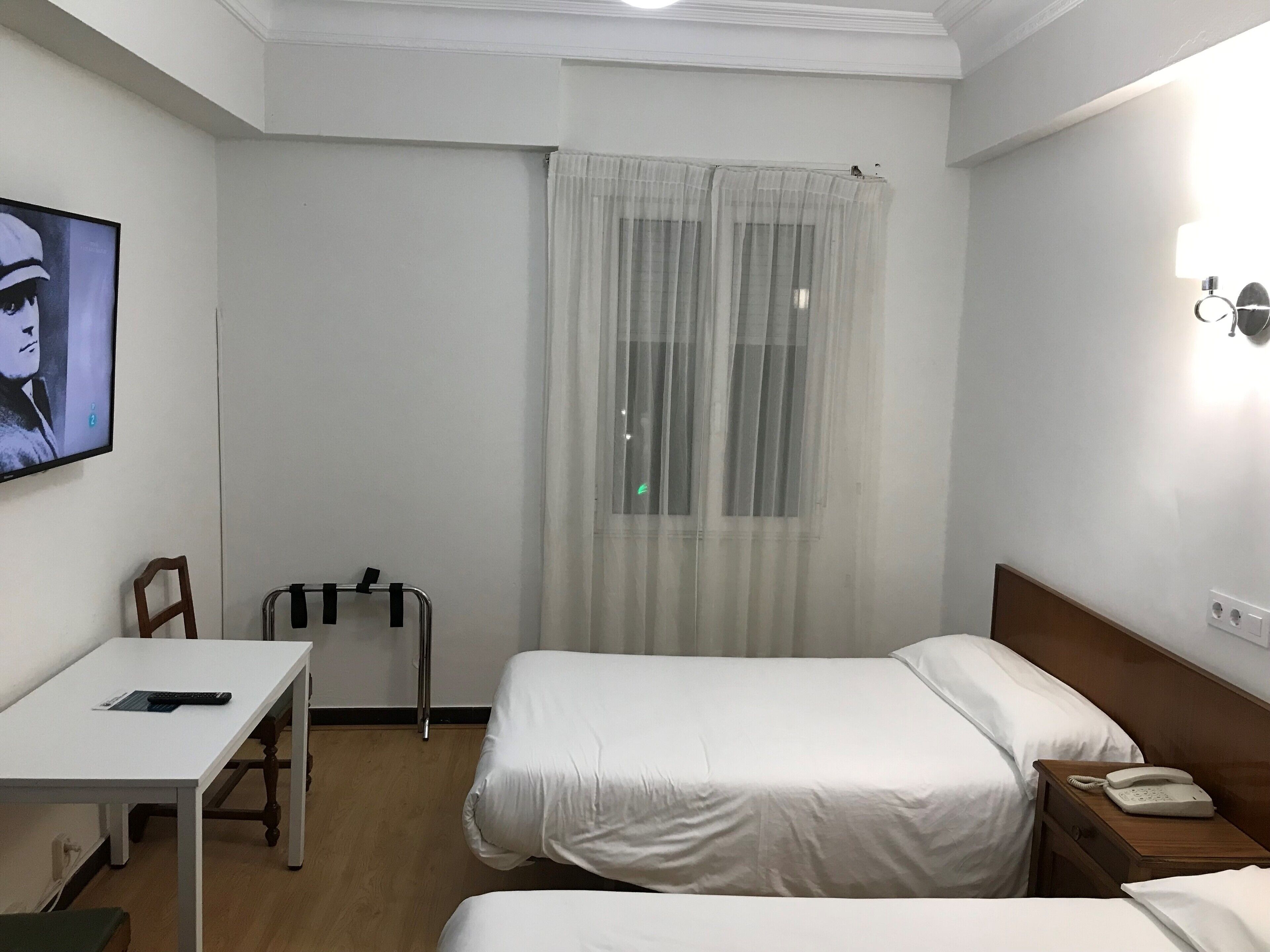 A hotel room with two beds, a desk with a chair, a bedside table with a phone, a window with sheer curtains, and a painting on the wall.