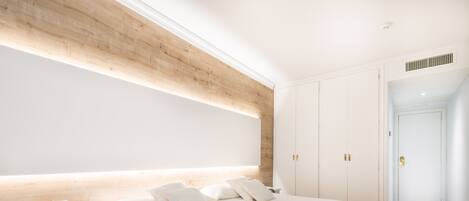 Un dormitorio moderno con una cama grande, cabecera de madera, paredes blancas y un banco empotrado con control remoto.