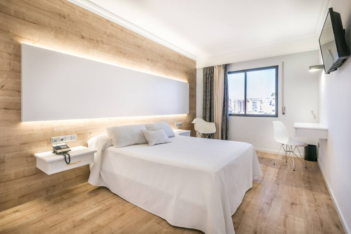 A modern hotel room with a large bed, bedside table, and a desk with a chair.