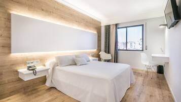 A modern hotel room with a large bed, bedside table, and a desk with a chair.