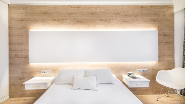 A modern bedroom with a large bed, bedside tables, a white chair, and a wooden wall. - Hotel Sansi Park (Lleida)