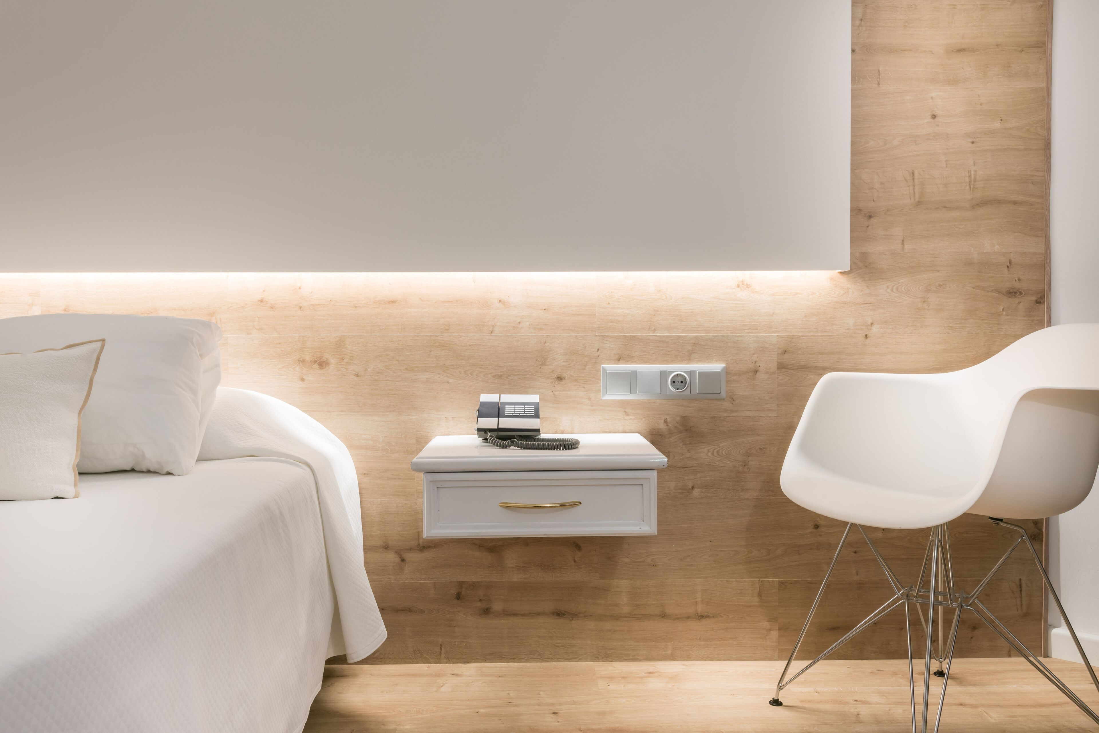 Un dormitorio moderno con cabecera de madera, una mesita de noche con lámpara, una silla blanca y una cama con ropa de cama blanca.