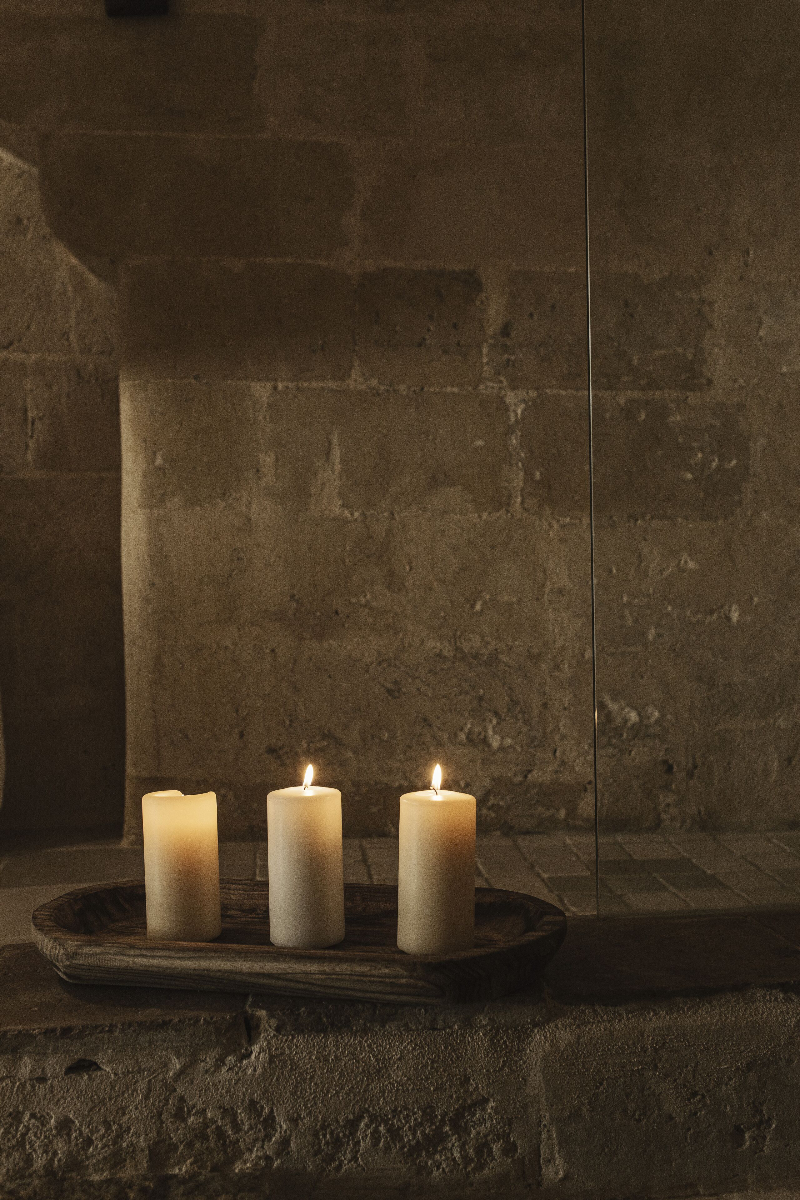 Três velas acesas sobre uma bandeja de madeira, com um muro de tijolos ao fundo.