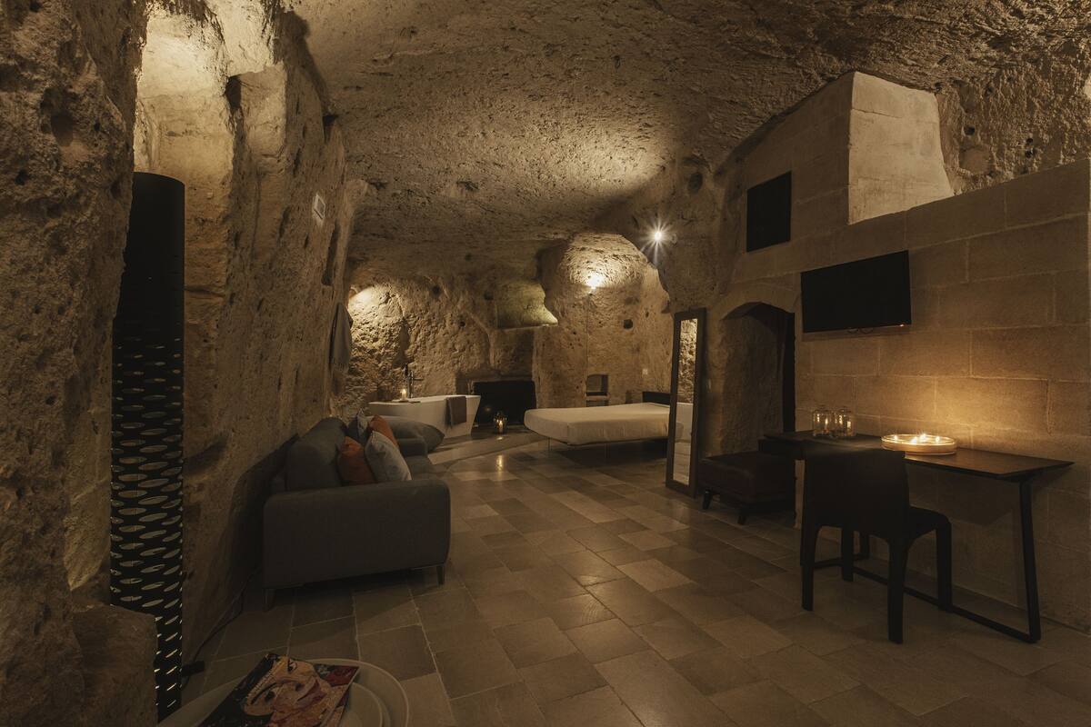 A rustic stone interior with a sofa, a bed, a table, and a fireplace.