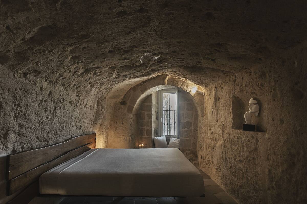 A bedroom with a stone ceiling, a wooden bed, a small window, and a statue on the wall.