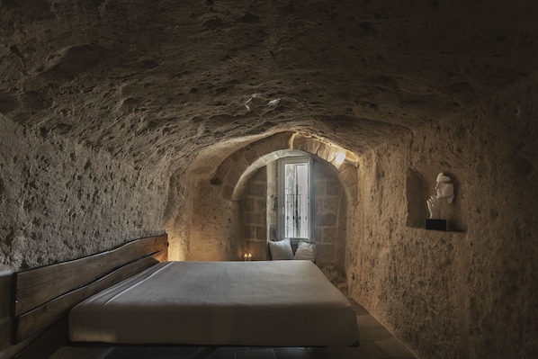 Un dormitorio con techo de piedra, una cama de madera, un ventanal pequeño y una estatua en la pared.