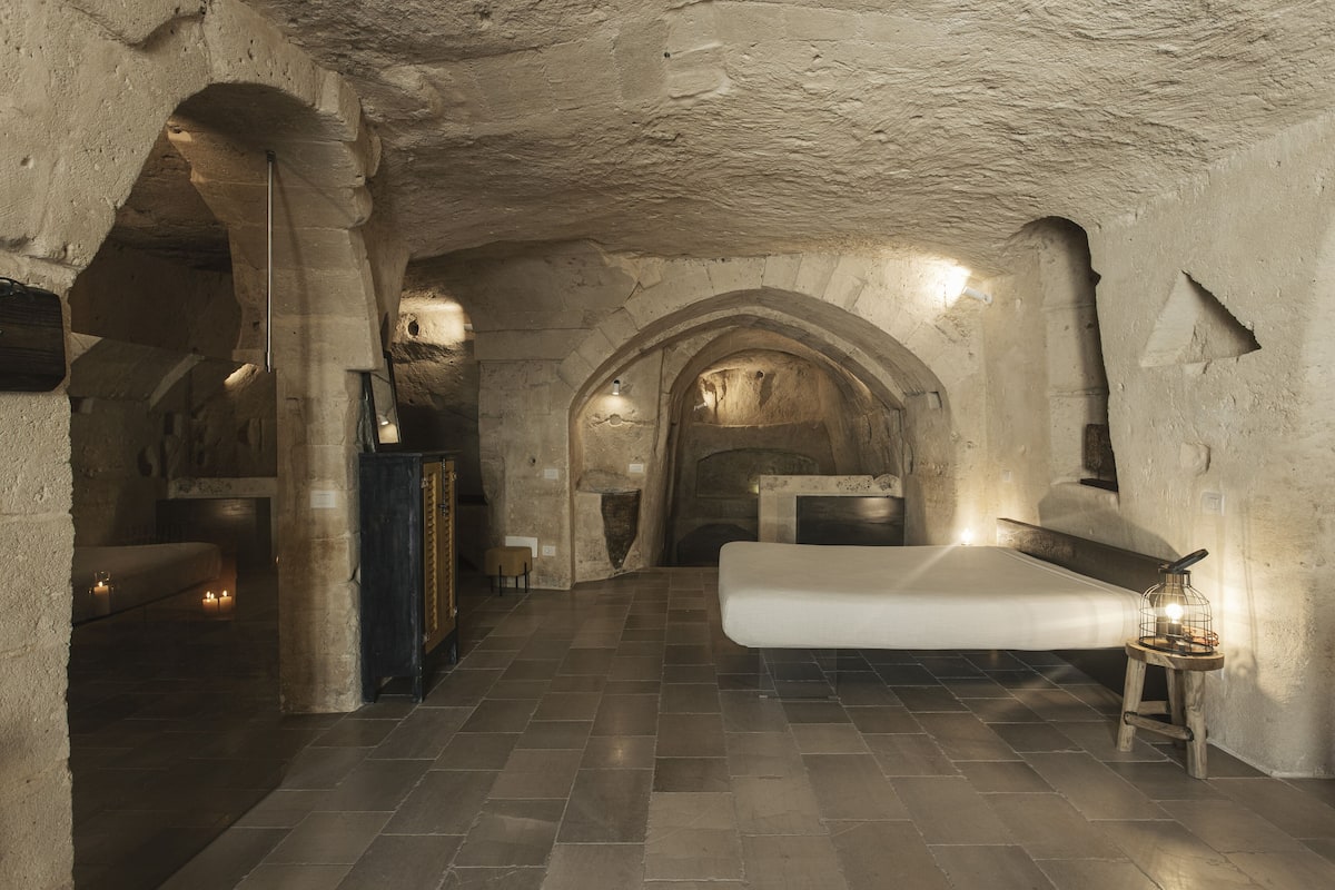 A room with a large bed, a wooden nightstand with a lamp, and a stone wall with an arched doorway.