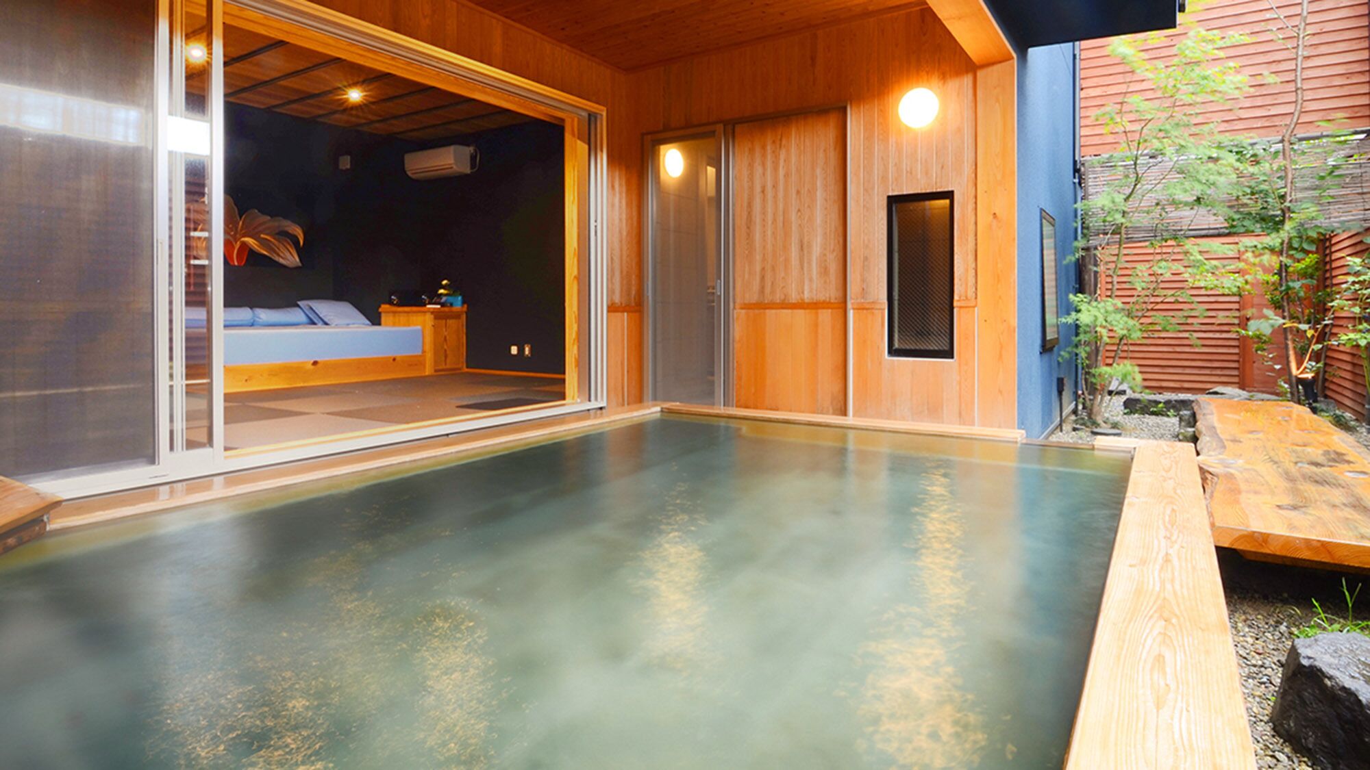 A traditional Japanese onsen with a wooden interior, a sliding glass door leading to a room with a bed, and a small garden area.