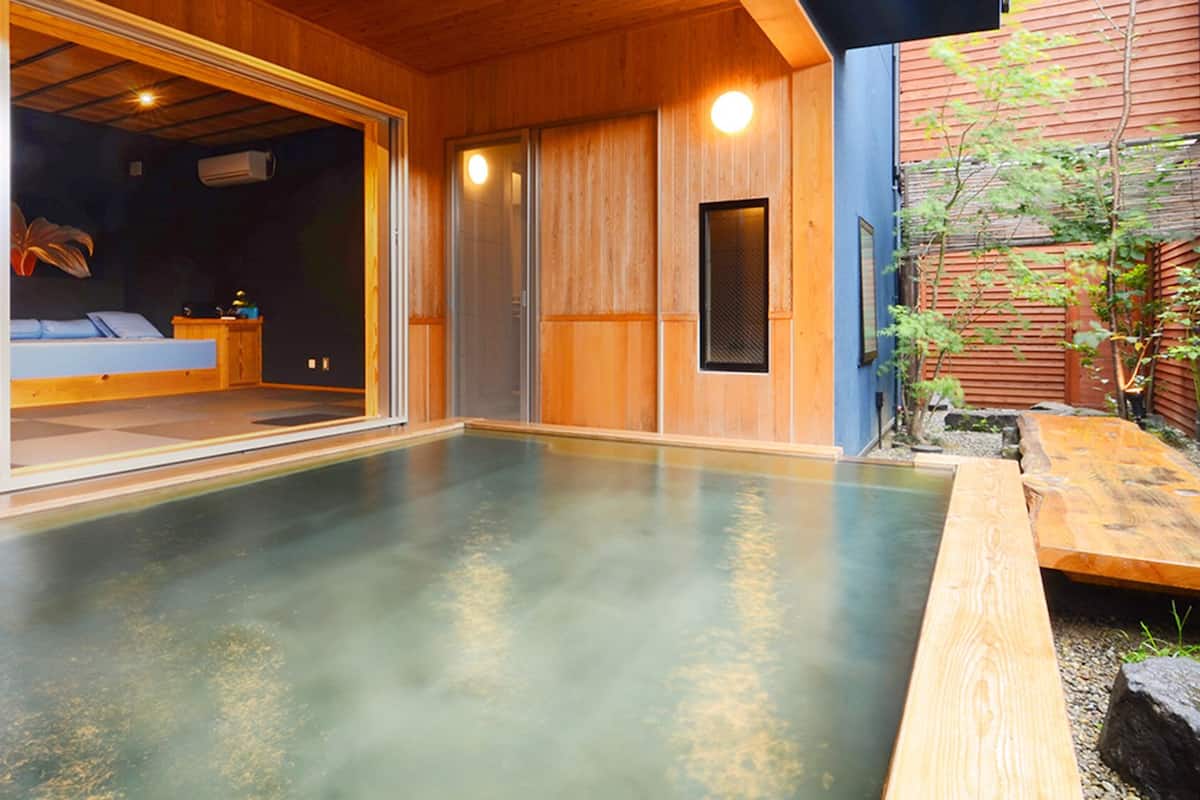 A traditional Japanese onsen with a wooden interior, a sliding glass door leading to a room with a bed, and a small garden area.