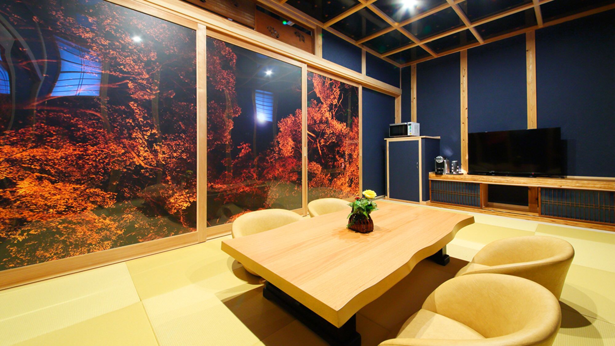 A conference room with a large table, chairs, a TV, and a view of autumn trees through large windows.