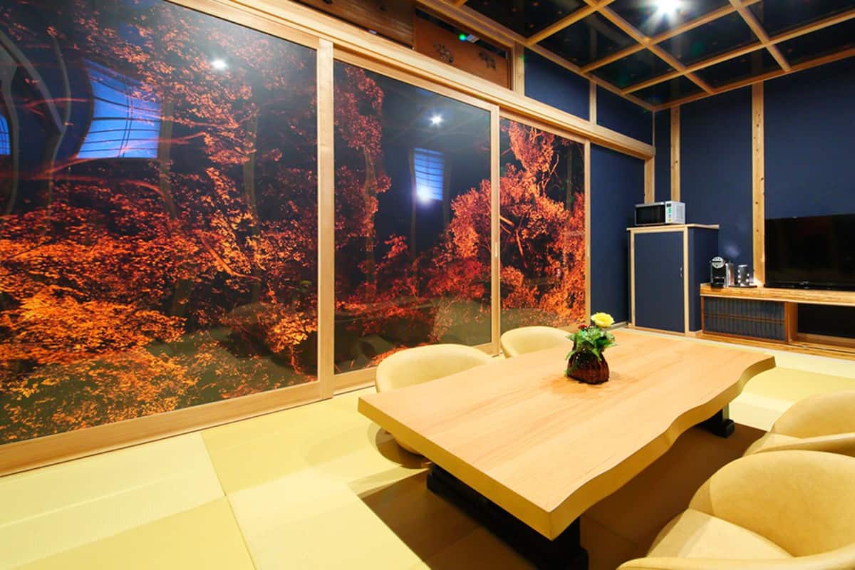 A conference room with a large table, chairs, a TV, and a view of autumn trees through large windows.