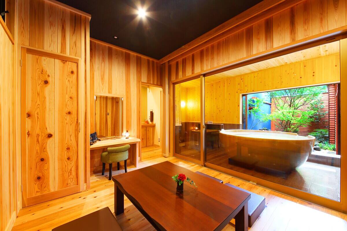 A wooden interior with a dining table, a bench, a small table with a vase, and a large bathtub visible through a glass door.