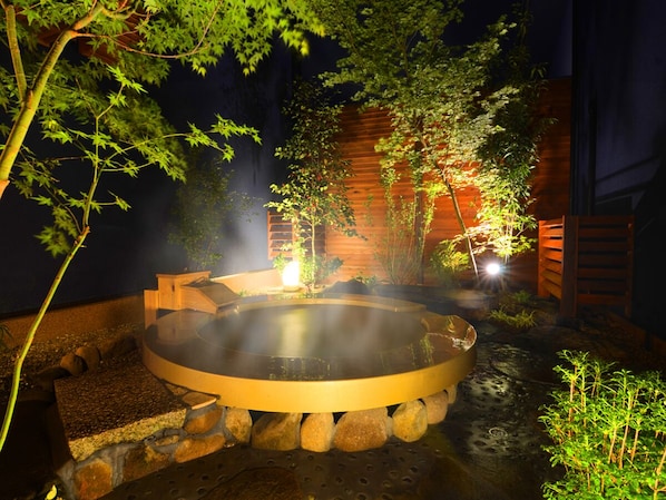 A hot spring bath surrounded by rocks and plants, with a wooden fence and a wooden structure in the background.