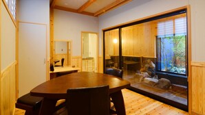 A dining area with a wooden table and chairs, a kitchen in the background, and a view of a garden through a large window. - Shosei no Yado Shinan (Beppu)