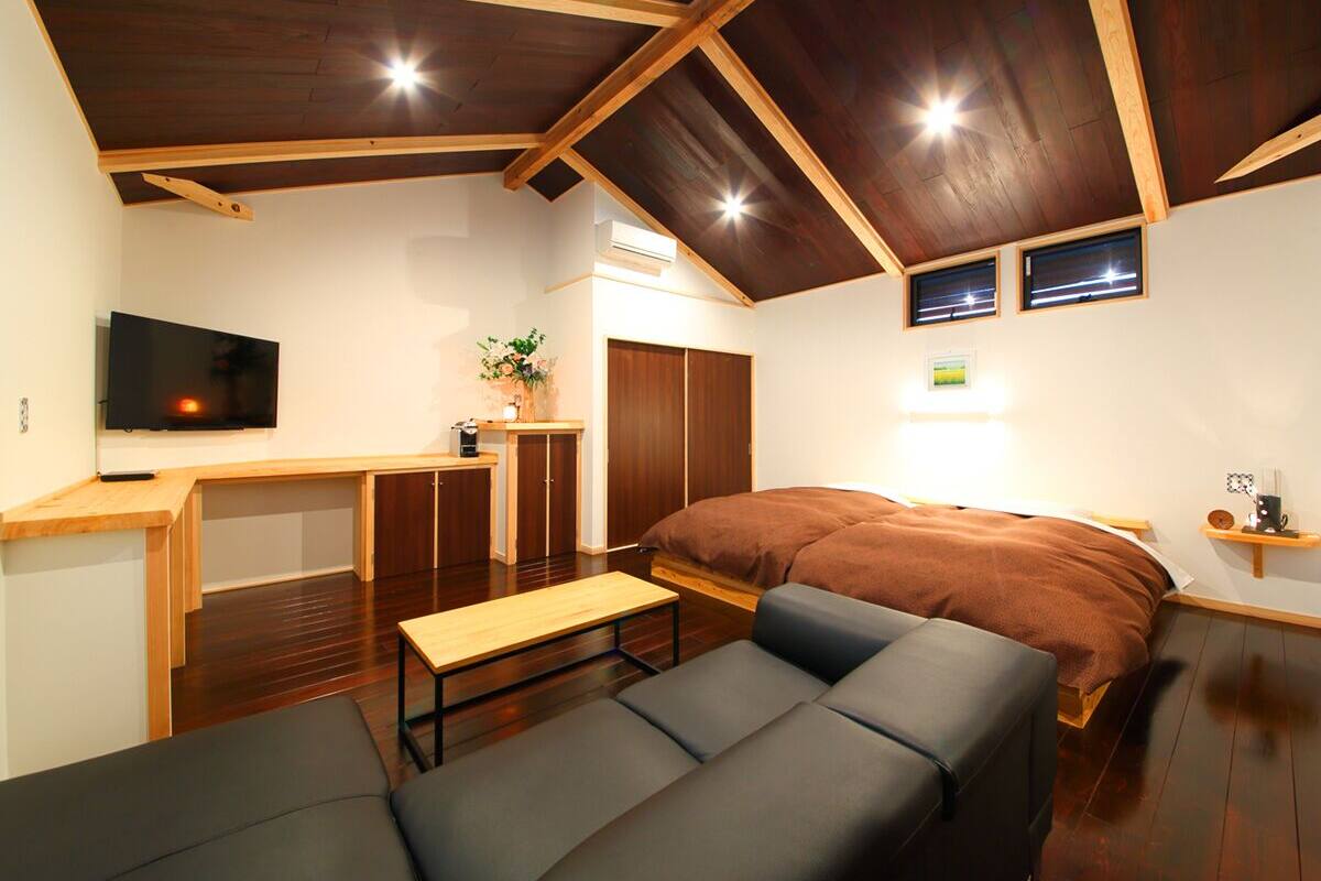 A compact room with a bed, a sofa, a small table, a TV, and a kitchenette area.