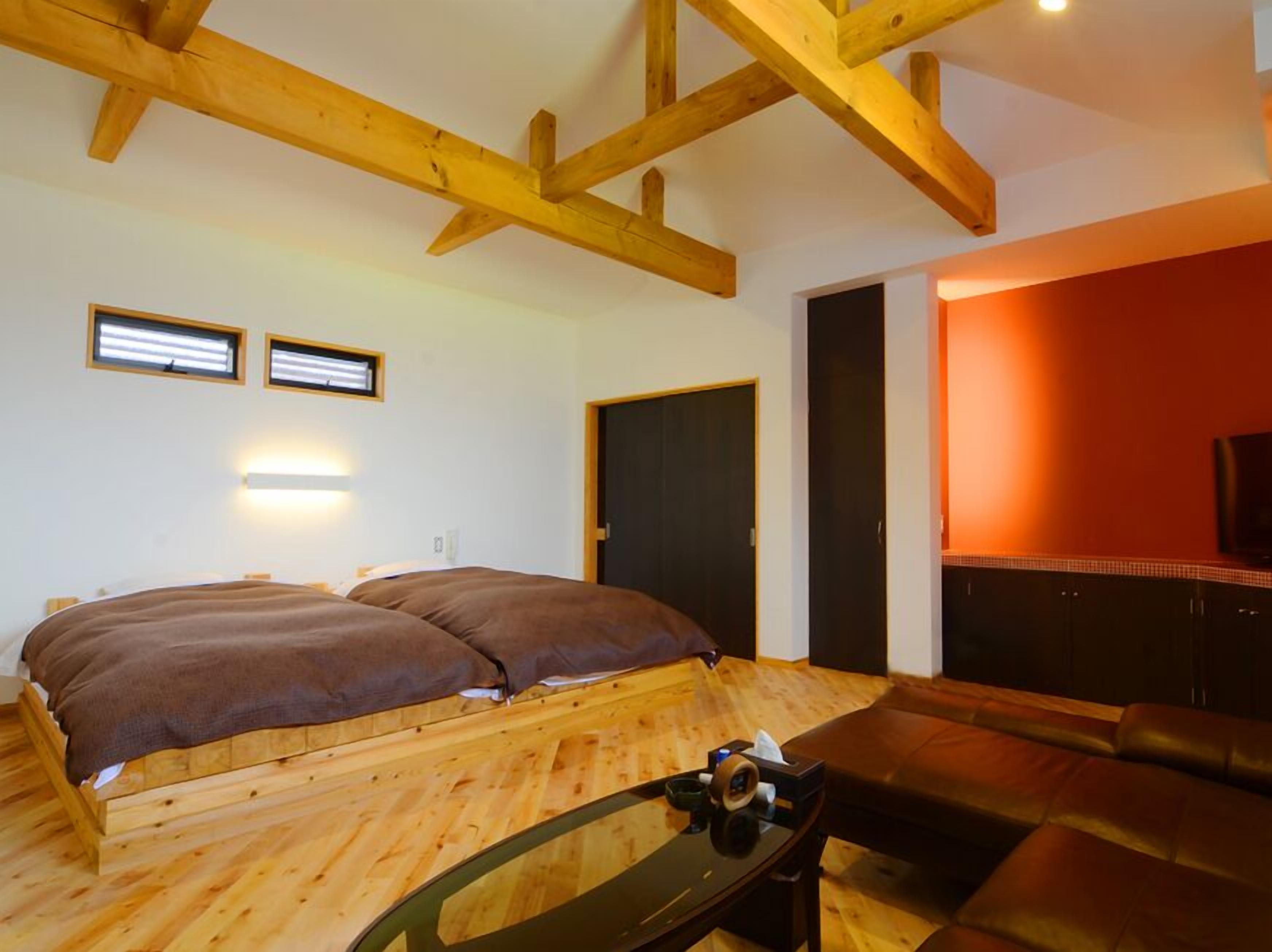 A room with a wooden floor, a bed with brown bedding, a glass table with items, a leather sofa, and a TV on a wall.