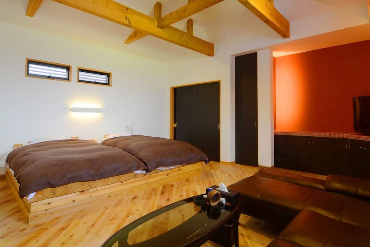 A room with a wooden floor, a bed with brown bedding, a glass table with items, a leather sofa, and a TV on a wall.