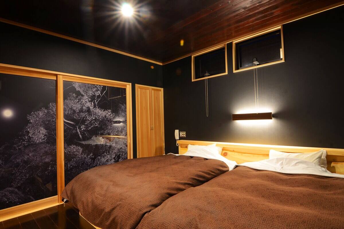 A bedroom with a large bed, a nightstand, a wall-mounted lamp, and a window with a view of trees.